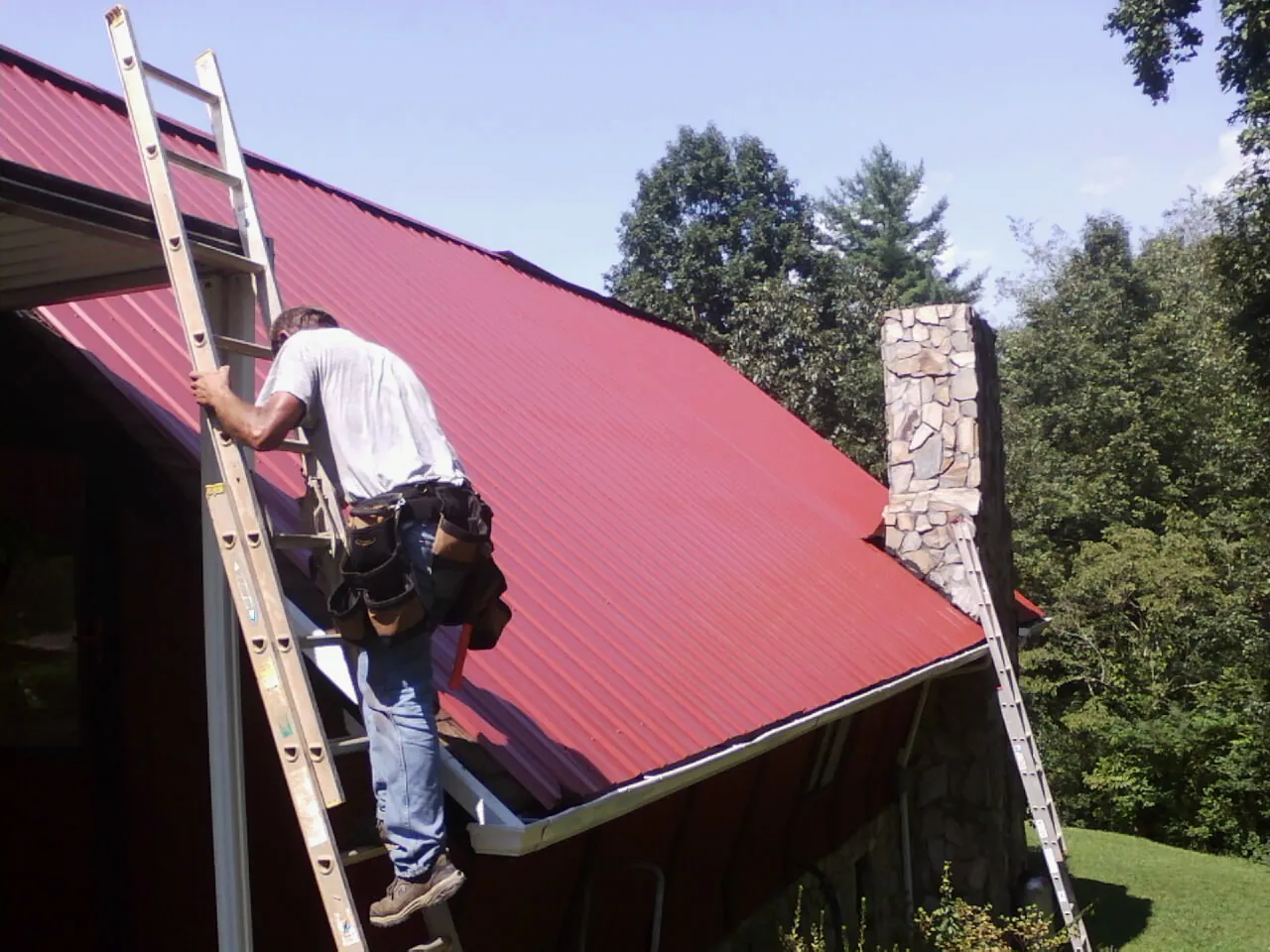 Roofer on a ladder beside a red metal roof and stone chimney — Soffit & Fascia Repair by Roofing Brothers in West Long Branch